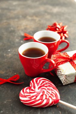 İki fincan sıcak espresso, kahve ya da tişört, güzel paketlenmiş hediye, kalp şeklinde beyaz-kırmızı lolipop ve desenli arka planda dekoratif fiyonklar. Sevgililer Günü romantik randevu konsepti, yakın çekim.