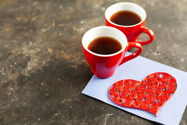 Two red cups of espresso or coffee, handmade heart postcards using quilling technique on a dark concrete background. Idea for romantic date for Valentines Day with drink and surprise. Selective focus