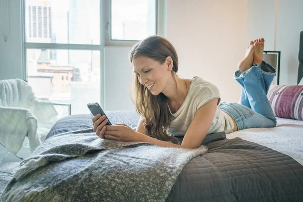 若い女性のベッドでの携帯電話のメッセージングを笑顔 — ストック写真