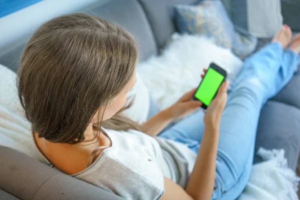 Young woman social networking on her mobile phone while lying on sofa in living room