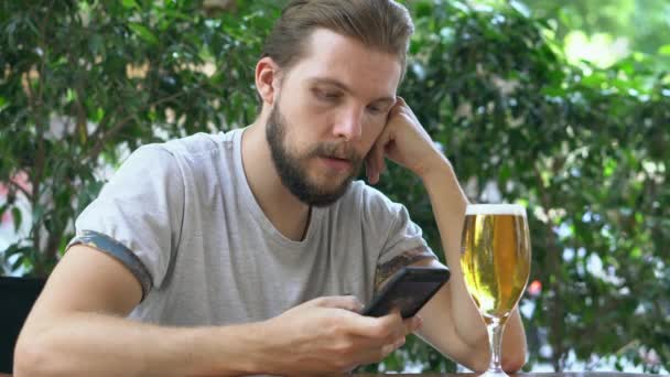 Молодой человек проверяет смартфон, пьет пиво — стоковое видео