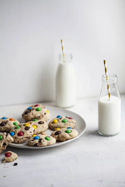 American cookies with chocolate chips and colored sweets, bottle with milk and straws on a gray background — Stock Photo, Image