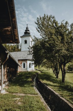 Eski Slovenya köyünün kıyısında kulesi olan eski beyaz bir kilise. Bahçedeki ağaçların altından akan dereler ve bahçedeki gölgeler. Güneşli sıcak yaz gününde dikey yönelimde.