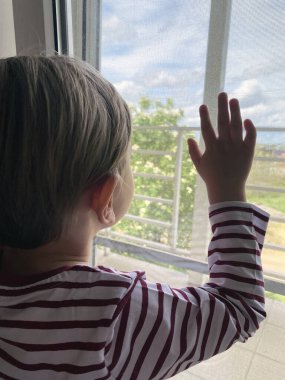 Çocuk evde kalır ve pencereden dışarı bakar. Oğlan arkadaşlarını ve sınıf arkadaşlarını özlüyor. Karantina sırasında, çocuklar izolasyon altında evde otururlar. Elini cama koydu.