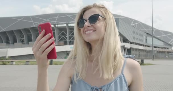 Hermosa chica en gafas de sol está sonriendo y haciendo selfie. Día soleado. Estadio en el fondo 4K — Vídeos de Stock