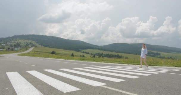 Una bella ragazza in occhiali da sole sorride e attraversa la strada su un passaggio pedonale. Giornata di sole. Belle montagne sullo sfondo 4K — Video Stock