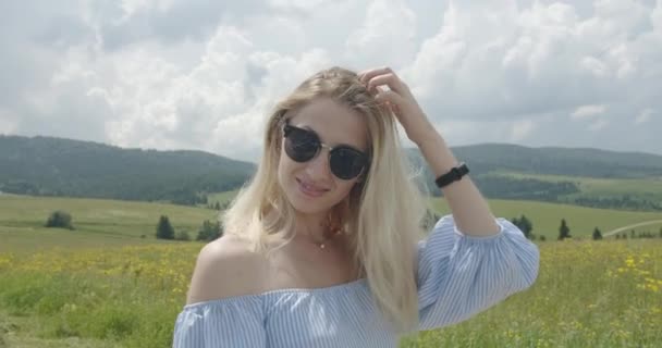 Hermosa chica en gafas de sol está sonriendo y alisando su cabello. Día soleado. Hermosas montañas en el fondo 4K — Vídeo de stock