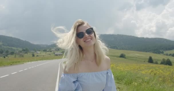 Hermosa chica en gafas de sol está sonriendo y alisando su cabello que se mueve por el viento. Día soleado. Hermosas montañas en el fondo 4K — Vídeo de stock