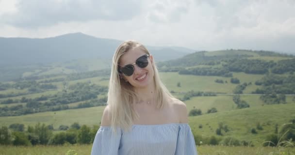 Hermosa chica en gafas de sol está sonriendo y mirando a las montañas. Día soleado. Hermosas montañas en el fondo 4K — Vídeo de stock