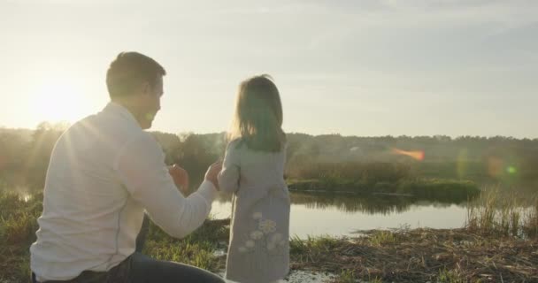 Un padre joven está viendo a su pequeña hija lanzar una ramita al lago. Familia en un paseo en el otoño. 4K — Vídeos de Stock