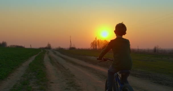 Un niño va en bicicleta por un sendero al atardecer. Filmando desde atrás. 4K — Vídeo de stock