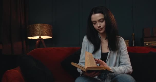 Das Mädchen liest gerade ein Buch und wechselt eine Seite, als es im Wohnzimmer sitzt. Abendstimmung. 4K — Stockvideo