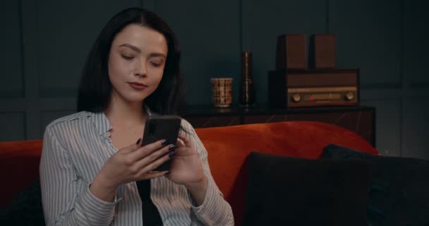 Ein schönes Mädchen entspannt sich auf dem Sofa und blättert Nachrichten auf ihrem Handy. Ein entspannter Abend nach Feierabend. 4K — Stockvideo