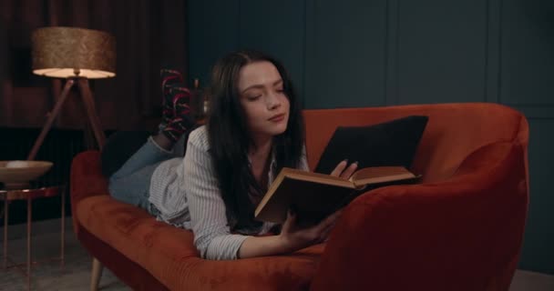 Молодая девушка лежит на диване и читает книгу. Добрый вечер после тяжелого дня. 4K — стоковое видео