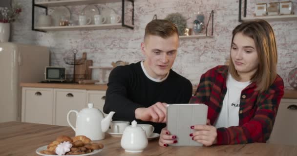 Un chico y una chica están viendo videos divertidos en la cocina en el desayuno. Hermosa mañana. 4K — Vídeo de stock