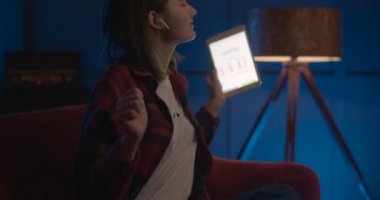 Kız kanepede oturuyor ve duygusal olarak tablete bakıyor. Oturma odasında rahatla. 4K