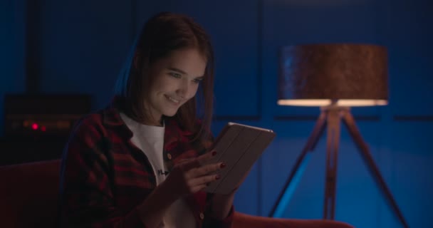 La chica está sentada en el sofá y sonriendo cuando mira la tableta. Relájate en la sala. 4K — Vídeos de Stock