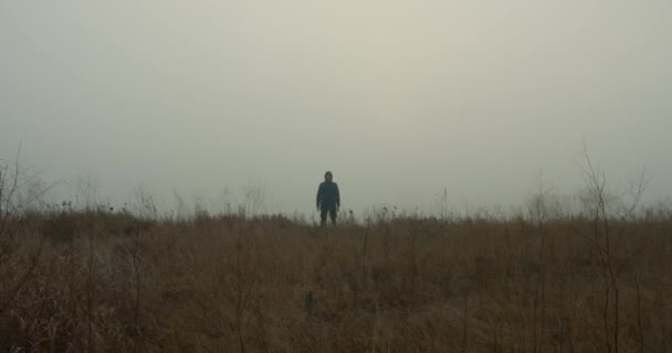 Mystical video. A boy in a hood stands on the edge of a field in the fog and looks at the camera. 4k — Stock Video