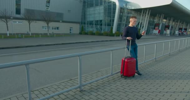 Хлопець пише смс по телефону, стоячи біля входу в аеропорт з валізою. Привіт, чекає літак. Сонячний день. 4K — стокове відео