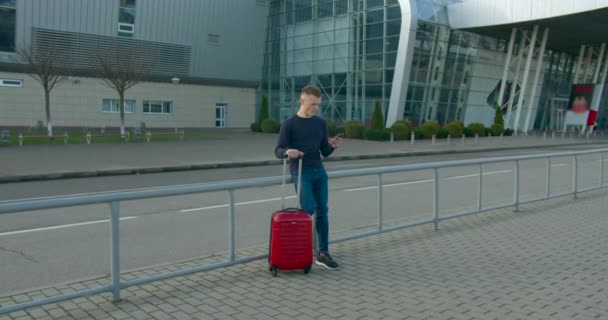 El tipo está escribiendo por teléfono y de pie en la entrada del aeropuerto con una maleta. Hermosa sonrisa. Esperando el avión. Día soleado. 4K — Vídeo de stock