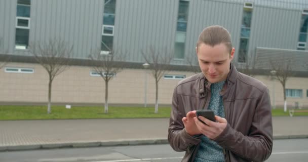 Un joven está enviando mensajes de texto por teléfono y de pie en la terminal del aeropuerto. Buen tiempo. 4K — Vídeo de stock