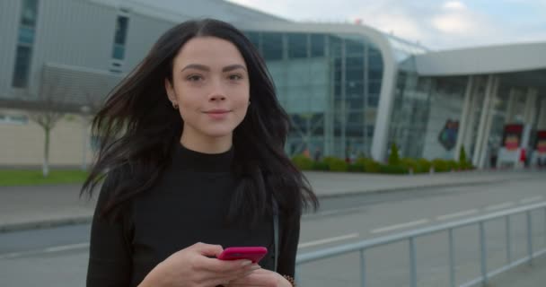 Κορίτσι πληκτρολογεί στο smartphone της και χαμογελά όταν στέκεται κοντά στο αεροδρόμιο. Τα μαλλιά της φτερουγίζουν στον άνεμο. Ταξίδι με αεροπλάνο. 4K — Αρχείο Βίντεο