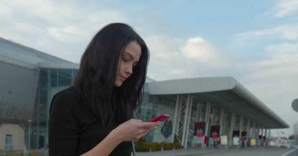 Όμορφη κοπέλα χαμογελάει και στέλνει μηνύματα και στέκεται κοντά στο αεροδρόμιο. 4K — Αρχείο Βίντεο