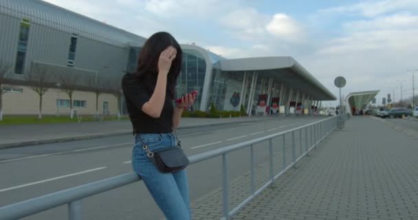 Hermosa chica está sonriendo y mensajes de texto en el teléfono cuando está de pie cerca del aeropuerto. Se está alisando el cabello. Viajar en avión. 4K — Vídeo de stock