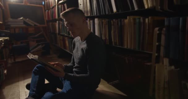 Egy fiatal fickó ül a padlón a könyvespolcok között, és egy könyvet lapoz. 4K — Stock videók
