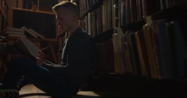 Un ragazzo è seduto sul pavimento tra le librerie e sfogliando un libro. 4K — Video Stock