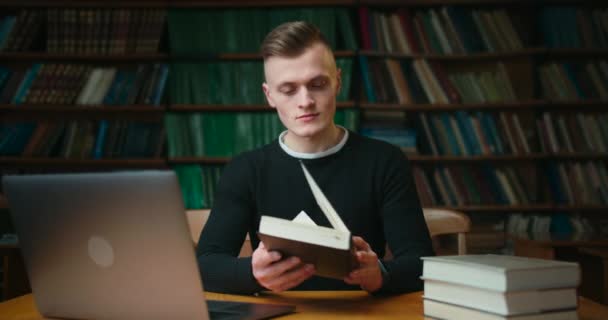 Młody człowiek zagląda do książki w czytelni biblioteki. Edukacja w bibliotece. 4K — Wideo stockowe