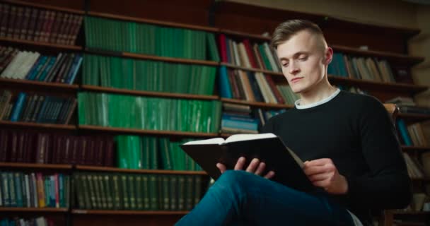 Il ragazzo sta lentamente guardando attraverso il libro che lo interessava. Il ragazzo è seduto sullo sfondo delle librerie. 4k — Video Stock