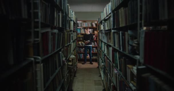 De man staat tussen de boekenplanken in het licht van het raam en leest een boek. 4k — Stockvideo