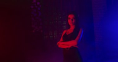 Güzel bir kız doğrudan kameraya bakıyor ve kollarını kavuşturuyor. Mavi ve kırmızı neon ışıklarla çekiliyor. 4k