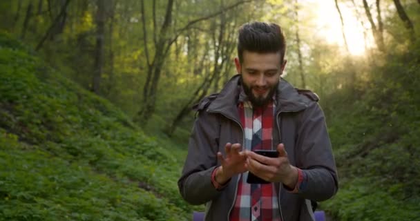 Der Kerl schaut auf die Karten auf dem Smartphone und geht durch den Wald. Wandern im Wald. 4K — Stockvideo