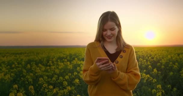这个女孩正在用电话发短信，日落时穿过田野。4K — 图库视频影像