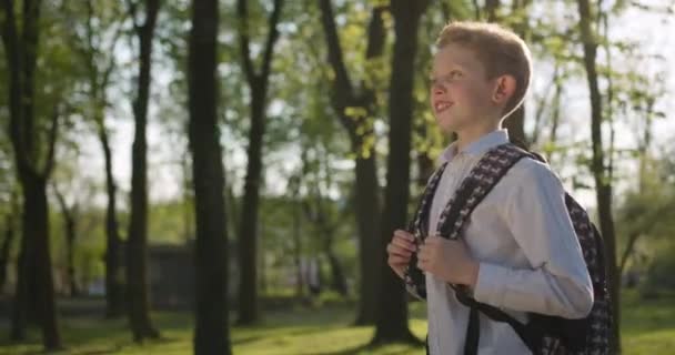 En leende pojke går genom parken med en ryggsäck. 4k — Stockvideo