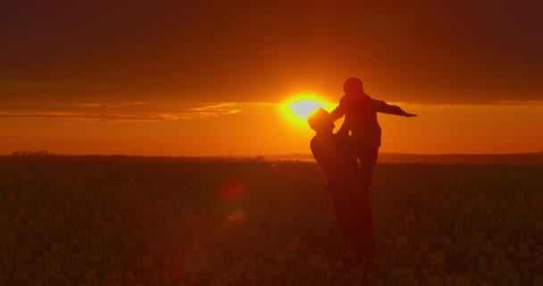 Un père tient son fils dans ses bras et le tord. Le champ au coucher du soleil. 4K — Video