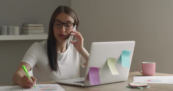 Das Mädchen mit der Brille sucht am Telefon nach Arbeitsfragen und macht sich Notizen. 4K — Stockvideo