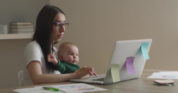 De moeder met bril werkt aan een laptop. De baby kijkt goed. 4K — Stockvideo