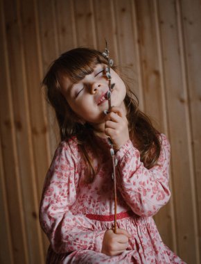 Güzel, mutlu, gülümseyen beyaz tenli, uzun kahverengi saçlı, kahverengi gözlü, söğüt dallı, pembe elbiseli, ahşap arka planlı bir kız.