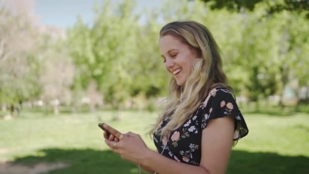 Веселая привлекательная блондинка, стоящая в парке с наушниками в ушах с помощью мобильного телефона улыбается и счастлив — стоковое видео