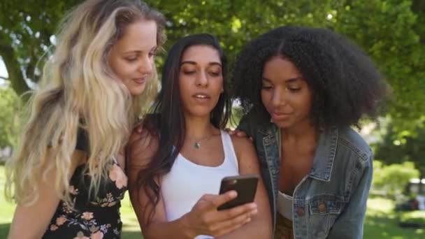 Щасливі різноманітні молоді друзі дивляться контент на екрані смартфона в парку друзі в парку за допомогою мобільного додатку — стокове відео