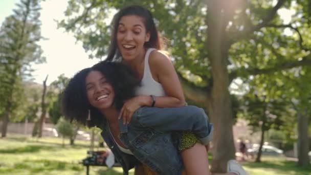 Счастливая молодая женщина перепрыгивает через своих подруг обратно и наслаждается катанием на спине в солнечный день в парке - африканская американская подруга дарит своему другу свинью назад — стоковое видео
