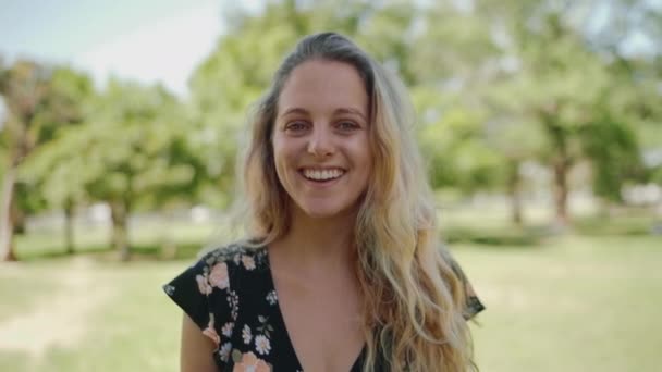 Retrato de una joven hermosa sonriente poniendo su mano en el pelo rubio mirando a la cámara en el parque sonriendo — Vídeos de Stock