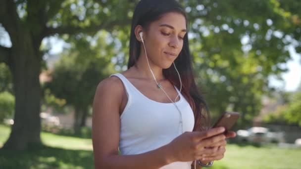 Cámara panorámica sobre la joven mujer escuchando música en los auriculares mensajes de texto en el teléfono inteligente en el parque - chica feliz en su teléfono en el parque en un día soleado — Vídeo de stock