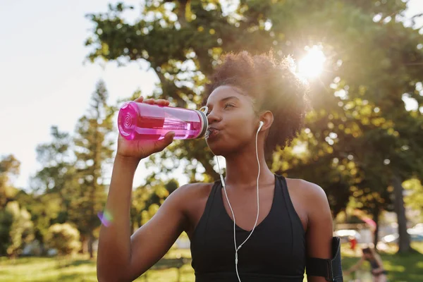 Fitness atleet jonge Afrikaanse Amerikaanse vrouw luisteren naar muziek op oortjes drinkwater in een herbruikbare fles water na het sporten uit te oefenen op zonnige dag in het park — Stockfoto