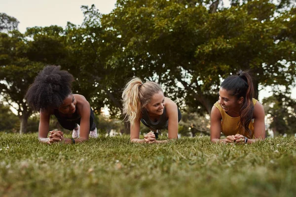 Tres jóvenes deportistas sonriendo mientras hacen ejercicios de tablón sobre hierba verde en el parque — Foto de Stock
