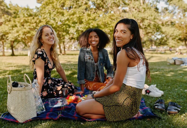 Senyum multirasial teman-teman perempuan duduk bersama di atas selimut di atas rumput hijau dengan buah-buahan di taman melihat kamera — Stok Foto