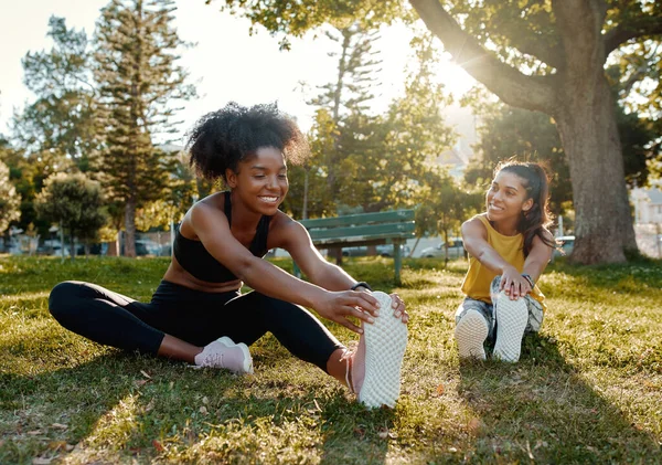 Giovani e diverse amiche sedute sull'erba verde che si allungano le gambe alla luce del sole mattutina al parco - amici diversi che si scaldano prima di fare esercizio di gruppo — Foto Stock